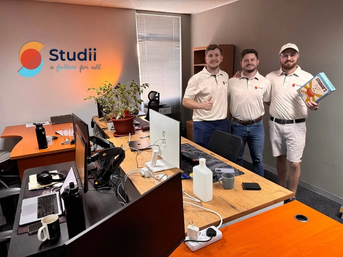 The three Studii founders in Studii shirts, standing in their office beneath the Studii logo